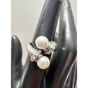 May include: A silver-toned ring featuring two faux pearls. Each pearl is accented with a band of clear crystals. The ring is displayed on a black surface, showcasing its design and details. A stylish piece of jewelry.