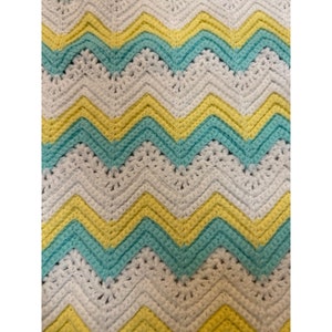 Op de afbeelding: Een gehaakt babydekentje met een chevron-patroon in wit, geel en lichtblauw.