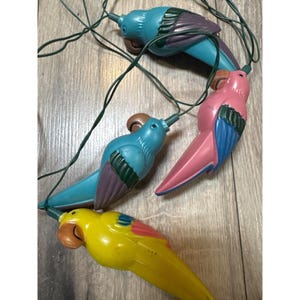 May include: A string of novelty parrot-shaped light covers connected by a dark green electrical cord. The parrots are molded in bright colors including yellow, pink, and two shades of blue, each featuring brown beaks and dark green wing details.