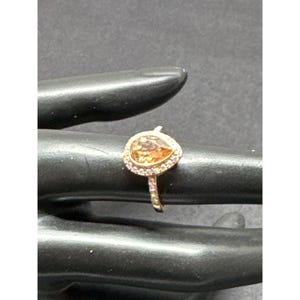 May include: A gold-toned ring featuring a pear-shaped, amber-colored gemstone surrounded by a halo of smaller, pinkish stones. The ring is set on a black surface, showcasing its intricate design and sparkle.