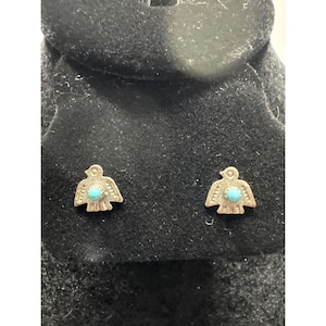 May include: A pair of silver-toned stud earrings, each shaped like a bird with outstretched wings. The bird's body features a small, round turquoise stone. The earrings are displayed against a black background.