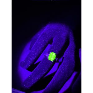 May include: A ring featuring a central emerald-cut gemstone that glows a vibrant neon green under ultraviolet light. The gemstone is set in a metallic band with smaller, less luminous stones on either side, creating a high-contrast visual effect.