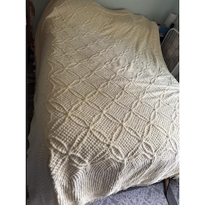 Könnte beinhalten: Cremefarbene, strukturierte Decke mit einem Rauten- und Kreismuster. Die Decke besteht aus einem weichen Material und ist über ein Bett drapiert. Das Design der Decke weist ein sich wiederholendes Muster aus Quadraten und Kreisen auf.