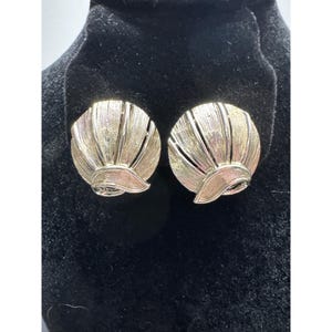 Puede incluir: Un par de pendientes dorados de clip. Cada pendiente presenta un diseño circular con un patrón en forma de abanico y un acento curvo en forma de hoja en la parte inferior. Los pendientes se exhiben sobre una superficie de terciopelo negro.