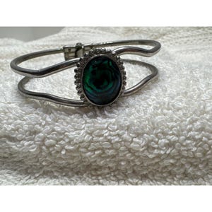 May include: Silver-tone hinged bracelet with an oval, iridescent, dark green stone. The stone is surrounded by a beaded border. The bracelet is resting on a white, textured surface.