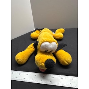 May include: A yellow plush toy of Pluto the dog from Disney. Pluto is lying on his belly with his tongue sticking out. He has black ears and a black nose.