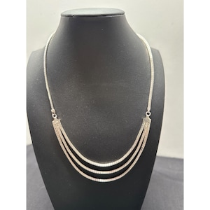 May include: A silver-toned necklace with a three-strand design. The necklace is displayed on a black mannequin. The strands are connected at the ends and hang in a gentle curve. The necklace is made of a shiny, reflective material.