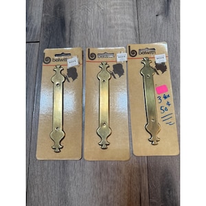 May include: Three brass-colored metal drawer pulls with decorative ends, each mounted on an individual tan cardboard backing card labeled with the Belwith brand logo. The pulls feature two screw holes and a smooth, elongated central bar design.