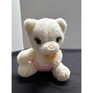 May include: A white plush kitten toy with green eyes, a pink nose, and a yellow pacifier in its mouth. The kitten wears a pink diaper. The toy is designed to resemble a baby animal.