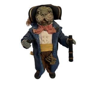 May include: A whimsical figurine of a sea creature dressed as a pirate captain. The figurine is wearing a blue coat, a red and white striped scarf, a yellow sash, and a black tricorn hat. The creature has a gray, textured face with a long snout and a mustache. The figurine is holding a telescope in one hand and a pipe in the other.