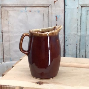 Pode incluir: Um jarro de cerâmica com acabamento castanho escuro brilhante e um esmalte de gotejamento bege mais claro à volta do rebordo. O jarro apresenta uma pega curva na lateral e um bico largo, exibindo uma superfície lisa e refletora em todo o corpo.