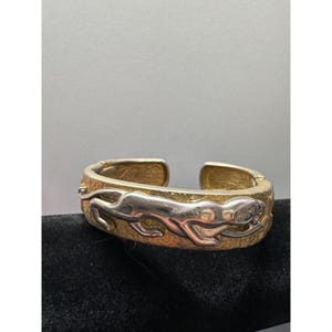 May include: Gold-tone metal cuff bracelet featuring a silver-tone cheetah design. The bracelet has an open design and is displayed on a black surface. The cheetah is in a running pose.