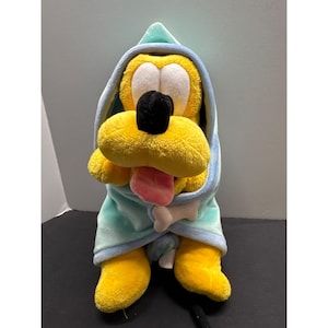 Puede incluir: Un perro de peluche amarillo que lleva una toalla con capucha azul con un diseño de hueso. El perro tiene la lengua rosa que sobresale.