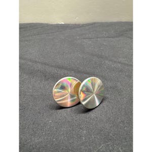 May include: Two circular, flat objects with a metallic, reflective surface that displays a holographic, rainbow-like sheen. The discs feature a radial pattern that catches the light, creating shifting colors of pink, green, and yellow across the faces.