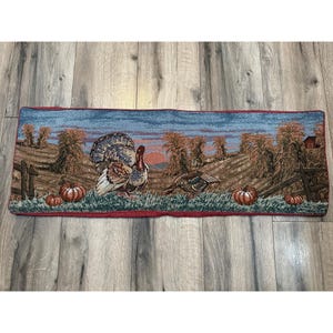 Pode incluir: Um tapete de porta com tema de Ação de Graças com uma borda azul e vermelha. O tapete apresenta um design de tapeçaria marrom, verde e azul com dois perus e abóboras.