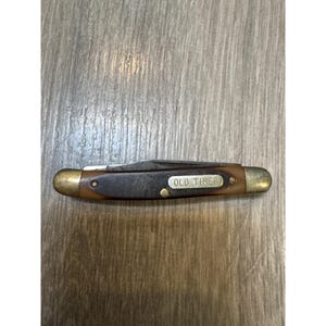 May include: A closed pocket knife with a dark brown textured handle and brass-colored metal bolsters at both ends. A rectangular metal inlay on the handle features the text OLD TIMER in embossed lettering. The blade is tucked inside the handle.
