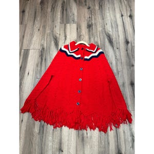 May include: Red knitted poncho with a white and blue trim and fringe. The poncho has a button closure.
