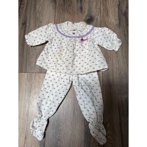 Puede incluir: Un conjunto de camiseta de manga larga y pantalón con pies en color blanco con un pequeño patrón repetitivo azul oscuro. La camiseta tiene un cuello con volantes, un borde morado en el pecho y un pequeño detalle gráfico rosa en el lado izquierdo.