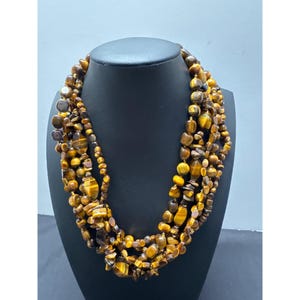 May include: A multi-strand necklace featuring tiger's eye beads in various shapes and sizes. The necklace has a warm color palette of browns, golds, and yellows. The beads are strung together, creating a layered effect. The necklace is displayed on a black mannequin.