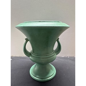 May include: A matte light green ceramic urn-style vase featuring a wide, flared rim, a rounded central body, and two curved handles on opposite sides. The piece stands on a circular pedestal base and has a subtle, speckled texture throughout.