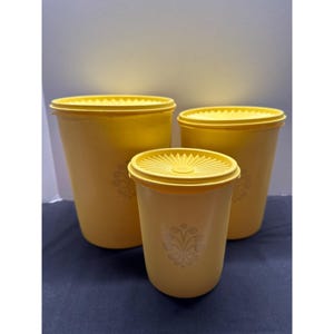 Peut inclure: Un ensemble de trois boîtes de conservation en plastique jaune avec couvercles assortis. Chaque boîte présente une forme cylindrique et un motif floral décoratif imprimé en couleur claire sur le devant. Les couvercles ont un motif en rayons.