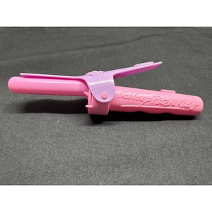 Può includere: Un arricciacapelli giocattolo in plastica rosa con una clip incernierata color viola chiaro e il testo in rilievo Cabbage Patch Kids sul manico. Il dispositivo presenta un cilindro e un meccanismo a molla per la clip come accessorio.