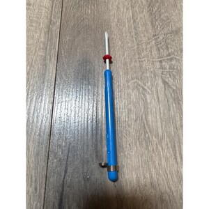 May include: A crochet hook featuring a bright blue ergonomic handle, a silver metal shaft, and a small red rubber ring positioned near the top. A silver metal spring coil is wrapped around the base of the blue handle, providing a textured grip.