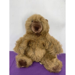 Puede incluir: Un oso de peluche marrón sentado en una superficie morada. El oso tiene una nariz marrón y ojos negros.