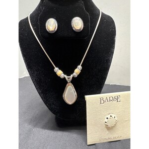 Pode incluir: Um colar de prata com um pingente em forma de pêra e brincos ovais a condizer. O colar tem pequenas contas de cor clara. Um pequeno cartão com a palavra "BARSE" e "STERLING SILVER" também é visível.