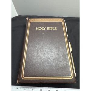 May include: A brown leather-bound Holy Bible with gold lettering and trim. The book is closed and has a textured surface. The title "HOLY BIBLE" is prominently displayed on the cover. The edges of the pages are visible.
