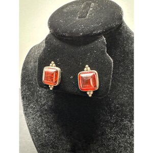May include: A pair of stud earrings featuring square-shaped, translucent orange-red stones set in silver-toned metal. Each earring is accented with three small silver spheres at the top and bottom edges, creating a symmetrical, geometric design.