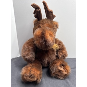 May include: A plush brown moose toy with a shaggy fur texture. The moose has large, dark brown antlers and a lighter brown snout with a small, tan-colored beard. The toy is sitting upright, with its legs extended.