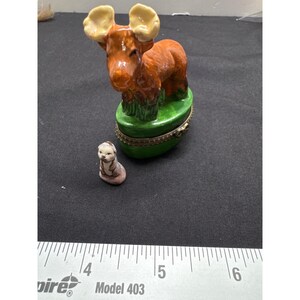 May include: A small ceramic figurine of a moose with brown fur and large antlers, standing on a green base. The moose is holding a small ceramic figurine of a white and brown animal.