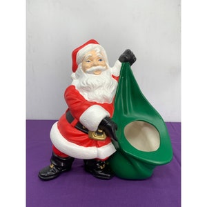 May include: A ceramic Santa Claus figurine holding a green sack. Santa is wearing a red suit with white trim and a white beard. The sack has a hole in the top, suggesting it is a planter.