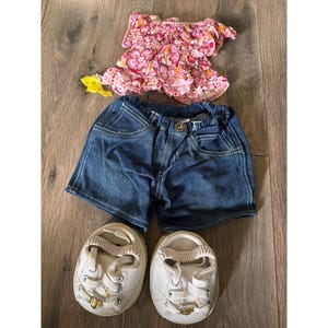 Op de afbeelding: Een poppenoutfit bestaande uit een top met korte mouwen met een roze en oranje bloemenpatroon, een donkerblauwe spijkerbroek met een elastische tailleband en twee witte sneakers met goudkleurige hartvormige bedels op de veters.