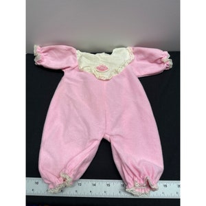 Könnte beinhalten: Rosaer Baby-Schlafanzug mit weißer Spitzenborte und einer rosa gestickten Blume auf der Brust. Der Schlafanzug hat lange Ärmel und einen gerafften Saum.