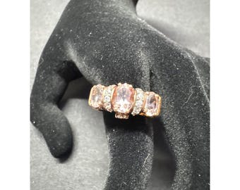 May include: A gold-toned ring featuring a central oval-cut pink gemstone flanked by two smaller, rectangular pink gemstones. The ring is further embellished with small, clear accent stones.