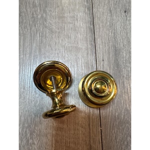 Könnte beinhalten: Zwei runde Schrankknöpfe mit poliertem Gold-Finish. Ein Knopf ist so positioniert, dass die kreisförmige Vorderseite und die zentrale Wölbung sichtbar sind, während der andere geneigt ist, um die Gewindeschraube an der Rückseite zu zeigen.