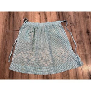 May include: A light blue and white gingham apron featuring a white cross-stitch floral pattern along the bottom hem and a single pocket on the upper left side. The apron includes two long fabric ties attached to the waistband for securing it.