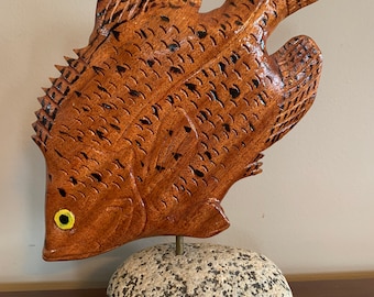 May include: A wooden carving of a fish with a brown stain and black details. The fish is mounted on a gray stone base.
