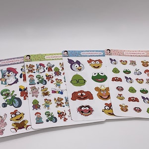 Peut inclure: Quatre planches d'autocollants avec divers personnages des Muppets. Les autocollants présentent des personnages comme Kermit la Grenouille, Miss Piggy, Fozzie Bear et d'autres. Chaque planche a un design différent, avec des illustrations colorées des Muppets dans diverses poses.