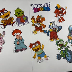 Könnte beinhalten: Eine Sammlung von Muppet Babies-Aufklebern mit bunten Zeichentrickfiguren wie Piggy, Kermit, Fozzie, Gonzo und Animal. Das Set enthält einen Muppet Babies-Logo-Aufkleber, verschiedene Charakterposen und Zubehör wie ein Dreirad, eine Staffelei und Bauklötze.