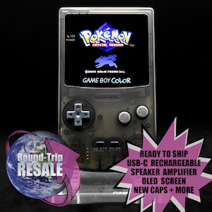 May include: A transparent Game Boy Color console displaying the Pokémon Crystal Version game. The screen shows the game's logo and a blue creature. The console has a directional pad, A and B buttons, and a start button. The text "Round-Trip RESALE" and "READY TO SHIP" are also visible.