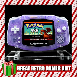 May include: A transparent purple Game Boy Advance handheld console displaying the "Pokémon FireRed Version" game. The console is on a clear stand, with the text "GAME BOY ADVANCE" visible. The image also includes the text "GREAT RETRO GAMER GIFT".