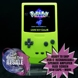 May include: A green Game Boy Color handheld gaming console with a black and white screen displaying the Pokemon Crystal Version game. The console has a black D-pad, A and B buttons, and a Nintendo logo.