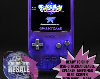 Könnte beinhalten: Eine violette Game Boy Color Handheld-Videospielkonsole mit einem durchsichtigen Kunststoffgehäuse. Der Bildschirm zeigt den Titel des Spiels Pokemon Crystal Version an. Die Konsole hat eine silberne Frontplatte mit Knöpfen und einem silbernen Steuerkreuz. Die Konsole steht auf einem durchsichtigen Ständer.