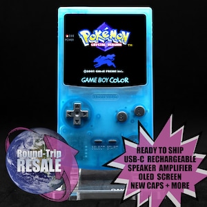 May include: A transparent blue Game Boy Color handheld game console displaying the "Pokémon Crystal Version" game. The screen shows the game's title and a blue silhouette. The console has gray buttons and the Nintendo logo.