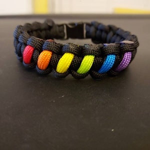 May include: A black paracord bracelet with rainbow colored stripes. The bracelet is woven with a knot pattern.