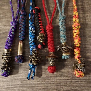 Könnte beinhalten: Sieben bunte Paracord-Schlüsselanhänger mit verschiedenen Metall-Anhängern. Die Anhänger umfassen einen Schädel, ein Dämonengesicht, einen Samurai, ein Monstergesicht, einen Oktopus und einen Helm.