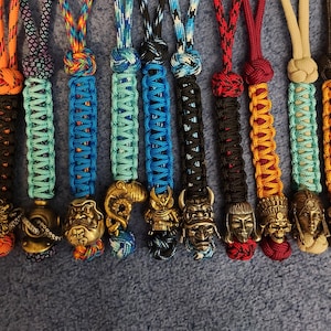 Op de afbeelding: Een verzameling handgemaakte lanyards met ingewikkelde gevlochten koorden in verschillende kleuren, waaronder oranje, blauw, zwart en rood. Elke lanyard heeft een unieke metalen bedel, zoals een duikhelm, octopus of samoerai.